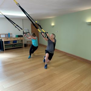 An older female and male lunging while holding onto TRX straps in a fitness studio with bamboo floor and green wall behind them
