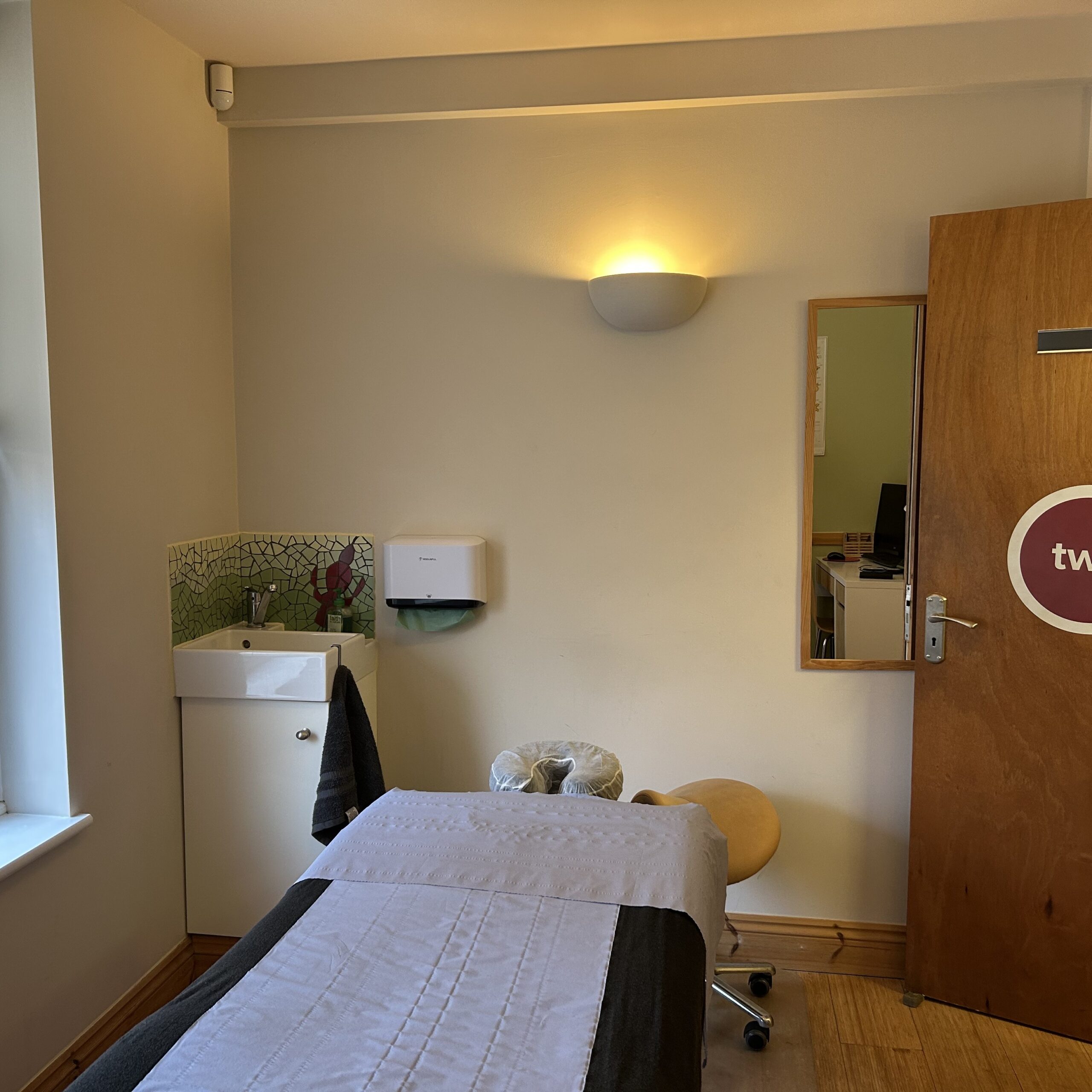 Treatment room showing basin, treatment couch and mirror. Light is on.