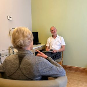 A practitioner is seated at a desk but facing a seated patient who has her back to us. He is smiling and holds a pen and pad in his lap.