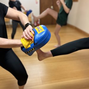 One figure holds a focus pad while another kicks it. Two people can be seen out of focus in the background doing the same move.